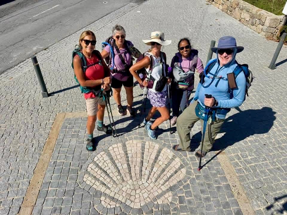 Five pilgrims with trekking poles pose on a cobbled road around a scallop-shell mosaic.