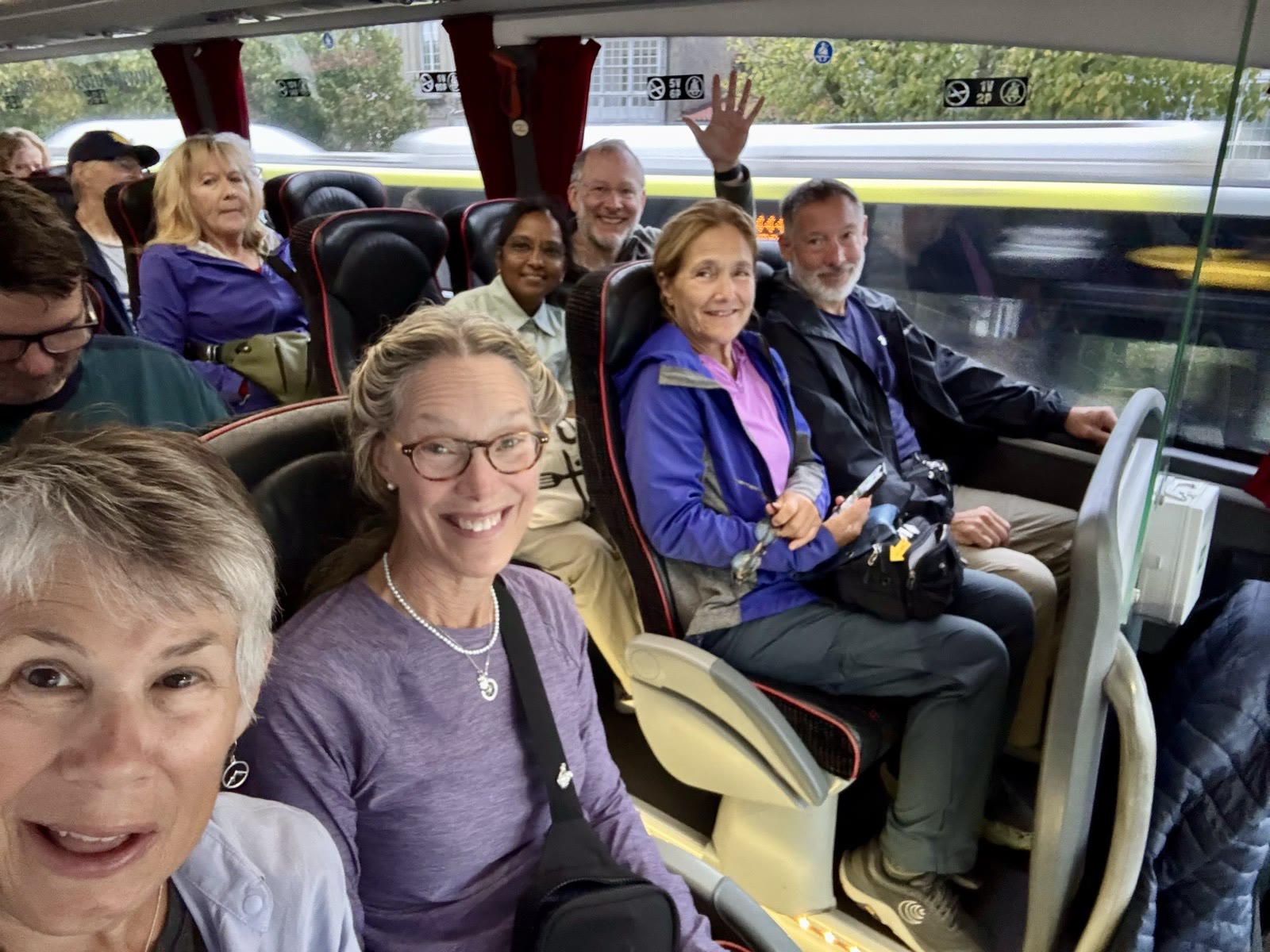 Smiling pilgrims in rain jackets seated on a tour bus, one in the back row waving.