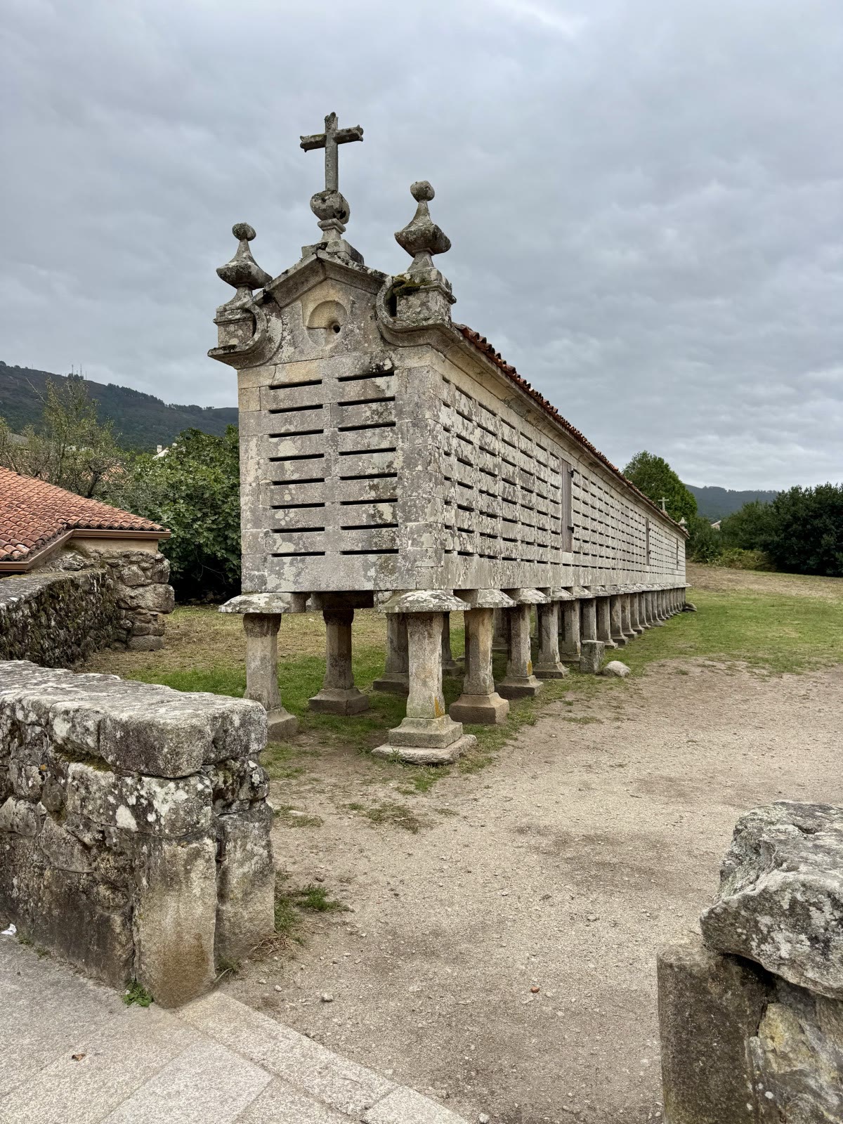 A long stone corn crib on stone pillars, topped with a cross and a red tile roof, beside a dirt path.