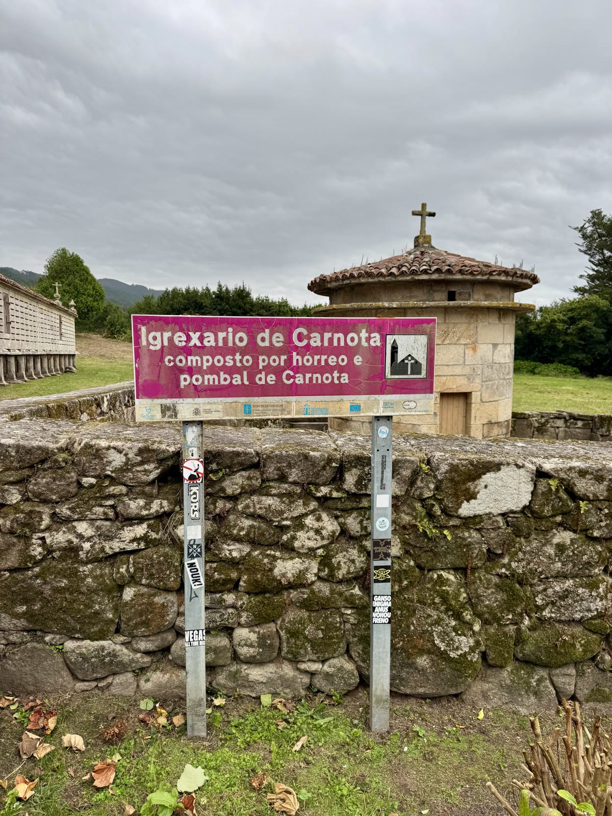 A pink sign reading Igrexario de Carnota, with a stone corn crib and a round stone dovecote behind it.