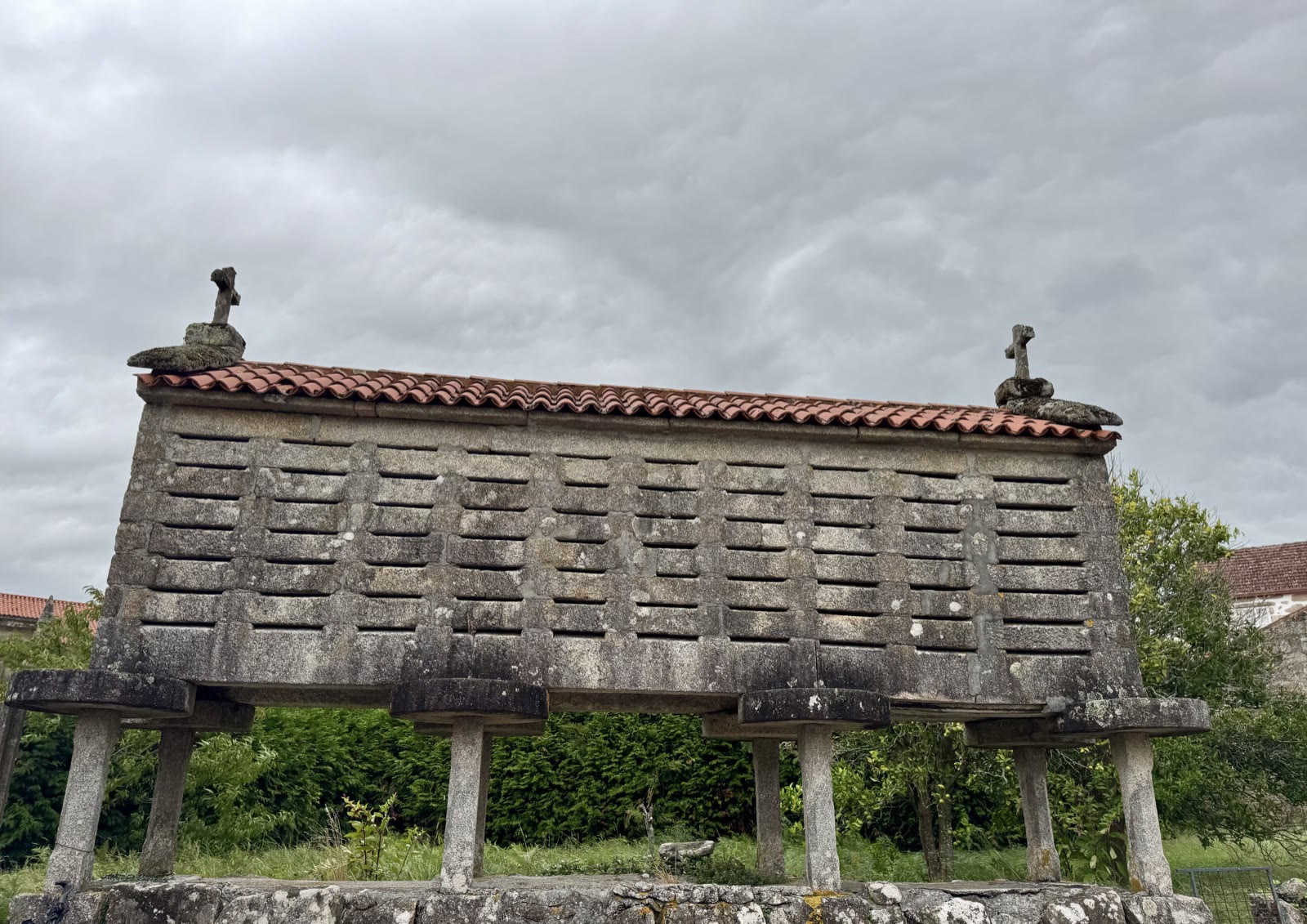A stone corn crib with a red tile roof and two stone crosses, raised on pillars against a stormy sky.