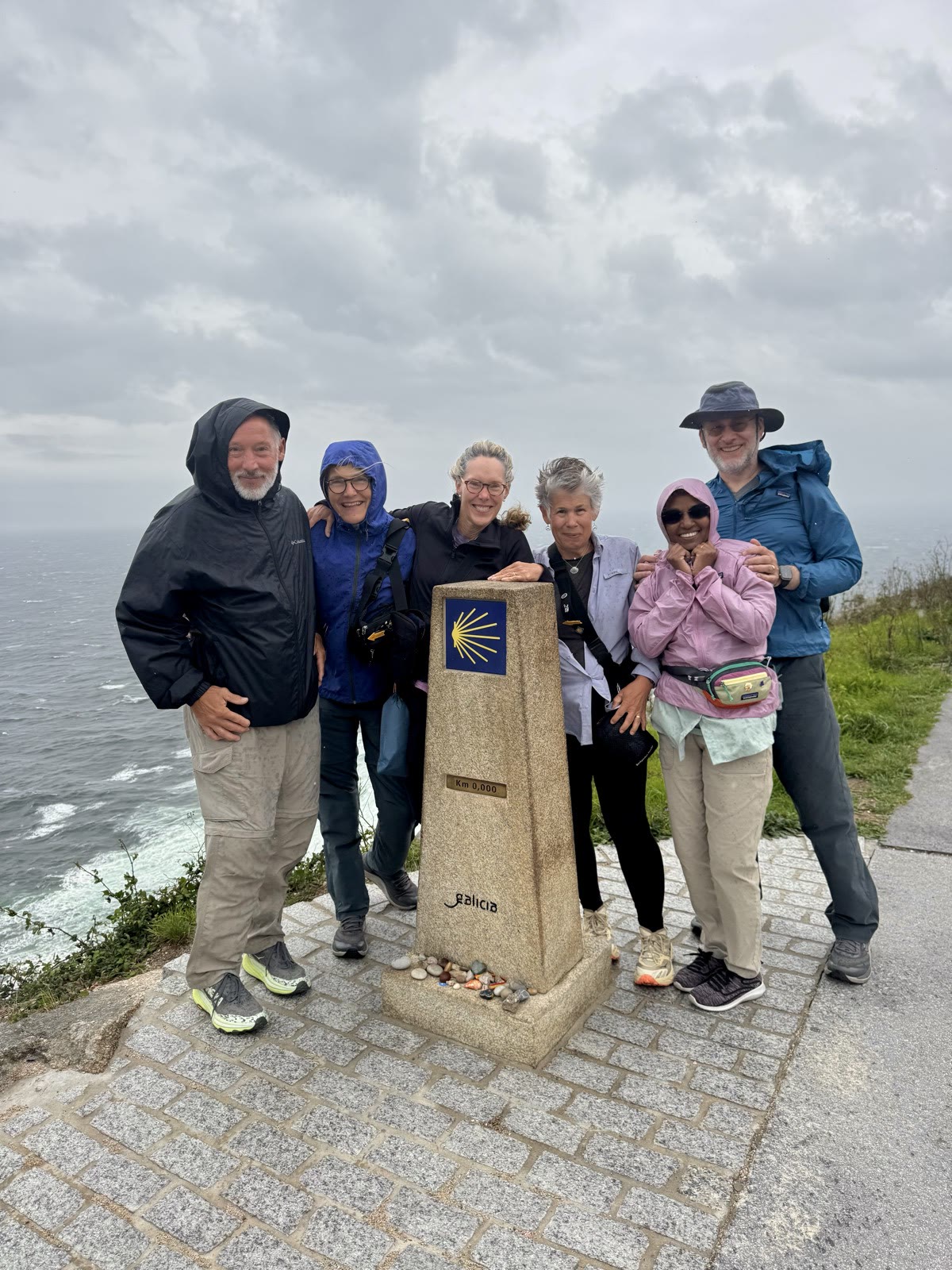 Six pilgrims in rain jackets pose in the wind beside the Km 0,000 stone marker on a cliff above the sea.