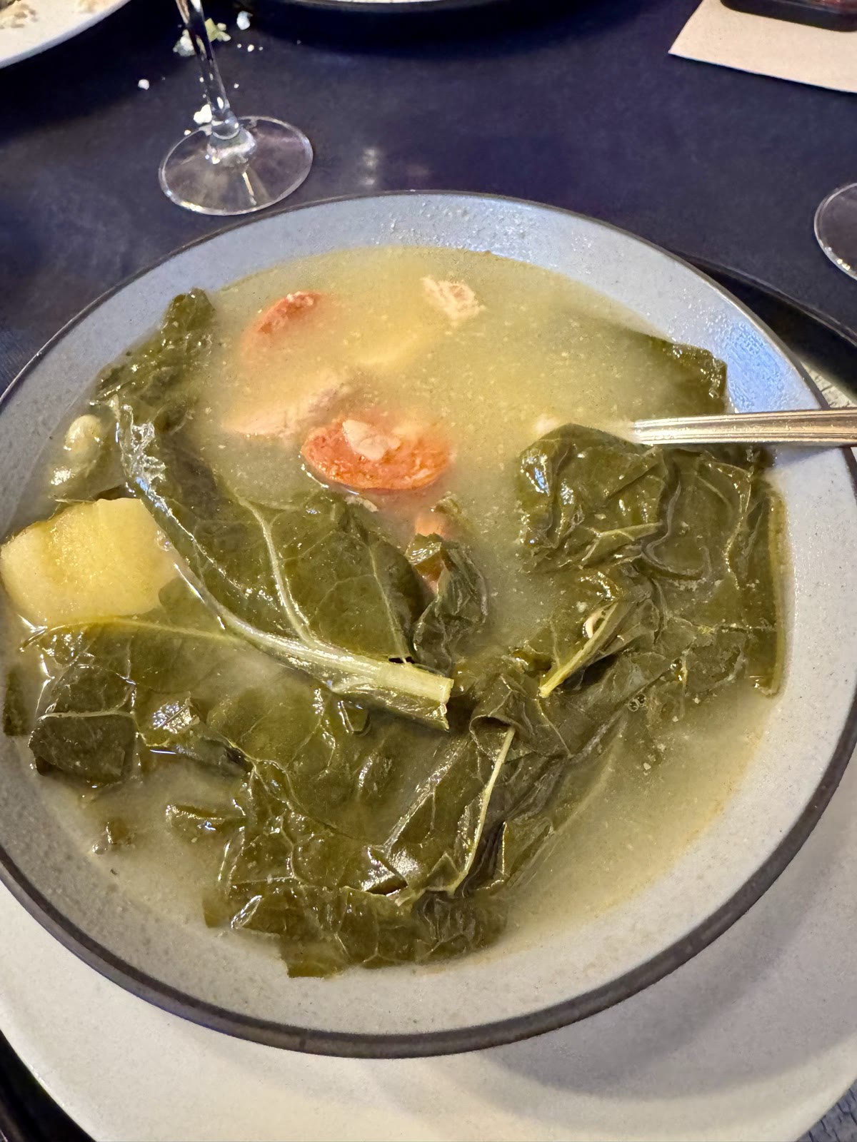 A bowl of broth with kale leaves, chorizo, pork, white beans, and potato.